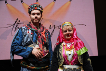 On the left stands a man, on the right a woman. They are dressed in traditional Turkish folk costumes. The man has a blue outfit with red and green accents and a colorful hat. The woman is wearing a pink and black outfit with ornaments and a pink headscarf. Behind them is a projection screen with a light pink background. A “thank you” sign appears in the background. Spotlights shine above the people. 