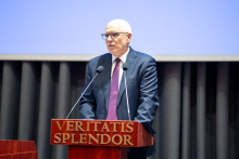 WUM Rector. A middle-aged man with glasses. He is wearing a dark suit, white shirt and reddish tie. He stands behind the rostrum and speaks. The rostrum is wooden and has the inscription “VERITATIS SPLENDOR” in gold. In the background is a blue multimedia screen hung with gray curtains.