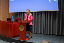 A middle-aged woman standing behind a lectern. The woman is wearing a pink jacket, black skirt and black shoes. The rostrum is made of dark wood and has the inscription “VERITATIS SPLENDOR” and a gold emblem on the front. A blue projection screen can be seen in the background. The screen shows white graphics in the upper right corner. To the left of the lectern is a desktop computer with a monitor positioned sideways to the viewer.  Translated with DeepL.com (free version)