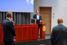 Three men in a conference room. Two of them have their backs turned to the lens, while the third stands behind the lectern and speaks. All are formally dressed. Behind the lectern is a multimedia screen with a blue background and a white logo in the upper right corner. The walls of the room are light and the floor is dark.