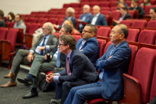 A group of people sitting in red chairs in an auditorium. Most of the people are dressed formally, in suits and ties. The background is blurred, but you can see more people sitting further away.