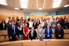 Group photo of about 35 people in the lecture hall. Some people are standing, some are sitting. They are smiling for the photo. Most people are dressed formally: in suits, dresses. Many have red badges around their necks. Wooden benches, bright walls and overhead lighting are visible in the background.