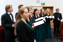 A group of people - choir members standing in a row inside the auditorium.They have their mouths open, singing.All are dressed in elegant, formal attire - suits, jackets or dresses. Most of them are holding open black briefcases or binders in which white sheets of paper are visible.