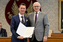 Dwóch mężczyzn w formalnych strojach, stojących obok siebie i uśmiechających się do zdjęcia. Mężczyzna po lewej stronie ubrany jest w granatowy garnitur, białą koszulę i krawat w paski. Trzyma otwartą teczkę z dokumentem. Mężczyzna po prawej stronie ma na sobie szary garnitur w kratę, białą koszulę i krawat w czerwono-niebieskie kropki. W tle znajduje się duże godło WUM z białym orłem na czerwonym tle. Obok herbu widoczny jest fragment obrazu w złotej ramie.