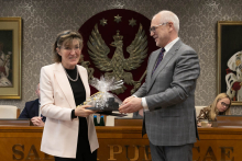 W przestronnej sali stoi obok siebie kobieta i mężczyzna. Kobieta stoi po lewej. Ma jasną marynarkę i czarne spodnie. Trzyma w rękach prezent, opakowany w przezroczystą folię z kokardą. Mężczyzna po prawej nosi szary garnitur. Obie osoby uśmiechają się. W tle widoczny jest czerwony herb z białym orłem oraz fragmenty dwóch obrazów na ścianie. Za mężczyzną i kobietą widać inne osoby, które siedzą.