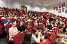 Duża sala wykładowa wypełniona studentami siedzącymi na czerwonych krzesłach. Studenci są zwróceni twarzami w stronę sceny, gdzie odbywa się symulacja rozprawy sądowej.