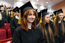 A group of graduates in the auditorium. They are all dressed in black academic togas and birettas with yellow grappling hooks. In the foreground on the left is a young girl with red hair, who smiles for the photo.