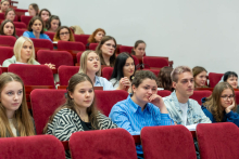 Grupa osób siedzących w auli w rzędach na czerwonych fotelach. Osoby na zdjęciu są różnej płci i wieku. Większość uczestników skupia uwagę na wydarzeniu odbywającym się poza kadrem – część z nich ma otwarte zeszyty lub notatniki. W tle widoczna jest biała, gładka ściana.
