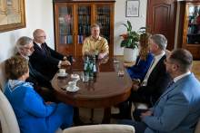 Seven people, rectoral authorities, deans and Prof. Craig C. Mello, in formal attire sit at an oval table in an elegant office. On the table are cups of coffee, bottles of water and glasses. In the background are wooden bookshelves and closed doors.