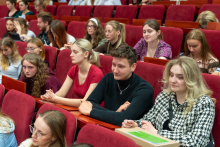 Grupa młodych osób siedzących w rzędach na czerwonych krzesłach. Na pierwszym planie widoczne są trzy osoby: kobieta w czerwonej bluzce z krótkim rękawem, mężczyzna w czarnym swetrze oraz kobieta w biało-czarnym, wzorzystym żakiecie. W dalszej części sali widać więcej uczestników, którzy siedzą i patrzą przed siebie lub przeglądają swoje notatki.