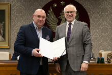 W przestronnej sali stoi obok siebie dwóch mężczyzn. Mężczyzna po lewej ma ciemny garnitur i koszulę w paski, a po prawej – szary garnitur i krawat w kropki. Mężczyzna po lewej stronie ubrany jest w ciemny garnitur i ciemny krawat, a mężczyzna po prawej w szary garnitur i krawat w kropki. Mężczyzna po lewej trzyma w rękach otwartą teczkę z dokumentem. W tle, na czerwonym tle, widoczny jest duży herb z białym orłem, a obok niego znajduje się obraz przedstawiający postać jednego z byłych rektorów uczelni.