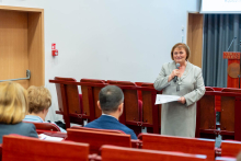 Kobieta w średnim wieku, ubrana w jasnoszary garnitur, stojącą w sali konferencyjnej i przemawiającą do publiczności. Kobieta trzyma mikrofon w prawej ręce i kartkę papieru w lewej. Przed nią, w kilku rzędach, siedzą słuchacze na czerwonych krzesłach z drewnianymi oparciami – widoczne są głównie tyły ich głów. W tle, po prawej stronie, znajduje się mównica oraz zasłonięte szare zasłony.