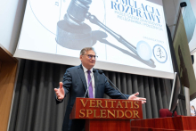Mężczyzna w garniturze stoi za mównicą z napisem "Veritatis Splendor". Mównica jest ustawiona przed dużym ekranem projekcyjnym, na którym wyświetlany jest obraz młotka sędziowskiego oraz stetoskopu. Na ekranie widnieje również tekst: "Symulacja rozprawy przed sądem zawodowym pielęgniarek i położnych" oraz logo organizatorów.