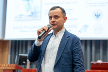 Mężczyzna w średnim wieku, ubrany w białą koszulę i niebieską marynarkę. Mężczyzna trzyma mikrofon w prawej ręce i mówi do niego. W tle widać ekran projekcyjny, na którym znajdują się logotypy różnych instytucji. 