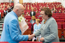 Aula w czasie konferencji. Na pierwszym planie, po lewej stronie, stoi mężczyzna ubrany w jasnoniebieską marynarkę, trzymający bukiet kwiatów owinięty w papier. Mężczyzna podaje rękę kobiecie stojącej naprzeciwko niego. Kobieta ubrana jest w szarą marynarkę i ma na szyi kolorowy naszyjnik. W tle widoczne są rzędy czerwonych, tapicerowanych krzeseł, na których siedzą uczestnicy wydarzenia. Większość miejsc jest zajęta, a osoby patrzą w kierunku sceny.
