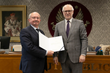 W przestronnej sali stoi obok siebie dwóch mężczyzn. Mężczyzna po lewej stronie ubrany jest w ciemny garnitur i ciemny krawat, a mężczyzna po prawej w szary garnitur i krawat w kropki. Mężczyzna po lewej trzyma w rękach otwartą teczkę z dokumentem. W tle, na czerwonym tle, widoczny jest duży herb z białym orłem, a obok niego znajduje się obraz przedstawiający postać jednego z byłych rektorów uczelni.