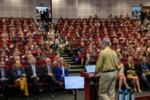 Aula z publicznością siedzącą na czerwonych krzesłach w czasie wykładu. Po prawej mężczyzna w jasnej koszuli i beżowych spodniach stoi przy podium z dwoma monitorami wyświetlającymi diagramy. W tle białe ściany z panelami akustycznymi i drzwi ewakuacyjne.