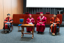 A group of people sitting on chairs in the middle of the auditorium. They are the rectoral and faculty authorities. They are all dressed in togas and academic birettas. In front of them stands a wooden table. Behind them you can see a section of a projection screen and gray curtains.