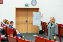 Na pierwszym planie, po prawej stronie, stoi kobieta w średnim wieku, ubrana w szary garnitur i trzymająca mikrofon. Kobieta zwrócona jest w stronę grupy osób siedzących po lewej stronie zdjęcia, co sugeruje, że przemawia lub prowadzi prezentację.  W tle, za kobietą, znajduje się tablica na stojaku z napisem „DZIEŃ PIELĘGNIAREK” oraz rysunkami przedstawiającymi pielęgniarkę i pielęgniarską czapkę z czerwonym krzyżem. 