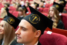 Close-up on the faces of the graduates facing the side of the photo, looking ahead. On their heads, the graduates are wearing black birettas with a gold embroidered logo of the Faculty of Pharmacy.