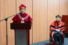 On the left is a female pro-rector, standing behind a wooden lectern, speaking into a microphone. She is dressed in a red academic toga with a decorative rector's chain and a red cap. A wooden wall is visible in the background. To the right of the photo sits another woman, she is wearing a toga. Behind them is a wooden wall.