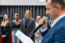 Cztery młode kobiety stojące w rzędzie na scenie auli. Wszystkie są zwrócone przodem do widowni i patrzą przed siebie. W tle znajduje się ciemna zasłona oraz fragment ekranu projekcyjnego.. Na pierwszym planie, po prawej stronie kadru, znajduje się mężczyzna trzymający mikrofon i kartkę papieru. 