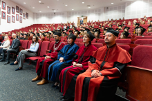 An auditorium with red armchairs where the participants of the diplomatatorium are seated. In the foreground sit the dean's authorities, dressed in official togas in colors representing the faculties: maroon, orange and blue.