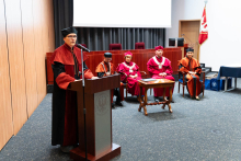 A group of people in the auditorium. In the foreground stands a woman, the associate dean for pharmacy, wearing an orange and black toga and black biretta. She stands behind a bronze lectern and speaks. Behind her, a group of people dressed in official academic togas sit on chairs. The togas are red or orange and black. Behind the people you can see a projection screen and gray curtains.