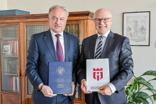 The two rectors stand side by side. The man on the left holds a blue folder with the words “Warsaw Medical University” and the university's emblem. The man on the right holds a gray briefcase with a red logo. In the background is a wooden bookcase with books. Above the bookcase hangs a painting. To the right of the picture stands a green potted plant.