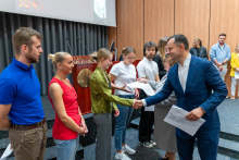 Wręczenie certyfikatów. Na scenie auli, w dwóch rzędach, stoi grupa młodych osób oczekujących na odbiór certyfikatów. Mężczyzna w niebieskim garniturze wręcza dokumenty. W tle widoczny jest drewniany pulpit oraz duży ekran projekcyjny. Pomieszczenie ma drewniane panele ścienne i ciemną podłogę.