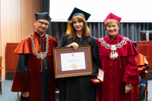 Three people posing for a photo. They are standing in the auditorium. The young woman in the middle, the graduate with the highest average, is dressed in a black toga and biretta. In her hands she holds a framed diploma. On the left stands the dean, on the right the pro-rector. Everyone is smiling.