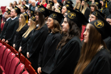 A group of young people. They are standing in rows, in front of them and behind them are red chairs. They are dressed in black togas and black birettas with a yellow faculty logo and a hoody. They are serious as they recite the words of the graduation oath.