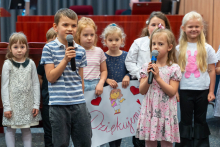 Grupa dzieci stojących na scenie auli. Dwoje dzieci na pierwszym planie trzyma mikrofony i śpiewa. W tle widoczne są inne dzieci, które trzymają duży, kolorowy plakat z rysunkiem kobiety oraz czerwonymi sercami. Na plakacie znajduje się napis „Dziękujemy!” wykonany czerwoną markerem. Dzieci ubrane są w różnorodne stroje.