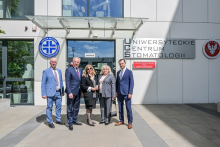 Five people in front of the entrance to the building. The individuals are elegantly dressed. The building has white walls and a large glass door. On the right side of the door a sign reads: “University Dental Center.”