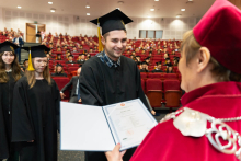 Young people, graduates, receiving diplomas from the pro-rector. In the foreground is a young man who smiles while receiving his diploma. The diploma being received is in an unfolded folder. The pro-rector is holding it with her left hand.