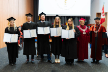 A group of 7 people. From the left stand the pharmacy graduates, three young women and two young men, dressed in togas. In their hands they hold folders with diplomas spread out. On the right stands the pro-rector and dean. They are all smiling. In the background, to the right, the WUM flag can be seen. A projection screen can also be seen behind the group.