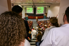 Lecture by Professor Craig C. Mello in the auditorium. The audience sits on chairs and steps, facing the speaker. In front of him are two screens with images resembling plant cells or biological tissues.