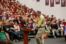 A man, Professor Craig C. Mello, wearing a yellow shirt, speaks at a lectern to a large audience in an auditorium. In the background are rows of red armchairs occupied by listeners and photos on the wall.