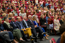 A large group of people are sitting in the auditorium on red chairs. Six people are visible in the front row, including a man in yellow pants and a woman in a blue suit.