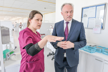 Two people standing in a bright room. The room resembles a dentist's office. The person on the left is wearing a maroon medical suit and a black shirt. The person on the right is wearing a dark suit, white shirt and maroon tie. Medical equipment can be seen in the background.