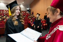 A young woman graduate, dressed in a toga and biretta, receives her diploma from the vice chancellor. The graduate has long curly hair. She smiles at the pro-rector. She is facing her. The pro-rector is also smiling. In the background, other graduates can be seen going to receive their diploma. 