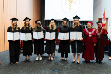 A group of 8 people. From the left stand pharmacy graduates, young women, dressed in togas.In their hands they hold folders with diplomas spread out.On the right stands the pro-rector and dean.They are all smiling.In the background, to the right, the WUM flag can be seen. A projection screen can also be seen behind the group. 