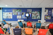 A group of people are sitting on colorful recliners in a conference room, watching a presentation on three screens. In the background are photos of operating rooms and the logo of Warsaw Medical University. The walls are decorated with posters of the university.