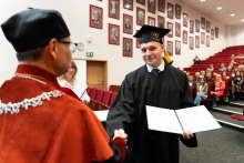 A young man, a graduate, wearing a biretta and toga, receives his diploma from the dean. The dean is facing the photo with his back, the graduate with his front. The graduate holds the unfolded diploma folder in his left hand. He is smiling. In the background you can see the rows of red armchairs where the guests of the ceremony are seated and the wall of the auditorium with framed portraits.