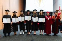 A group of 8 people. From the left stand the pharmacy graduates, one young man and 5 young women, dressed in togas. In their hands they hold folders with diplomas spread out. On the right stands the pro-rector and dean. They are all smiling. In the background, to the right, the WUM flag can be seen. A projection screen can also be seen behind the group.
