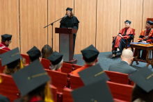 A woman dressed in a black toga and biretta stands in the middle of the auditorium behind the lectern and speaks into the microphone. In the foreground of the photo you can see the graduates looking at the speaker. You can see their upturned heads wearing birettas with clasps.