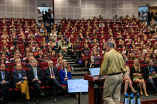 An auditorium with an audience sitting on red chairs during a lecture. On the right, a man in a light-colored shirt and beige pants stands at a podium with two monitors displaying diagrams. In the background are white walls with acoustic panels and an emergency exit door.