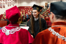 A young woman graduate, dressed in a toga and biretta, receives her diploma from the vice chancellor and dean. The graduate has long dark hair. She is smiling. The pro-rector and dean are shot from behind, their backs are visible, they have academic chains around their necks and birettas on their heads. In the background you can see red armchairs and the people sitting on them.