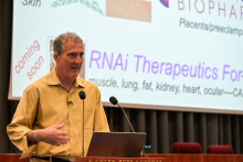 A man, prof. Craig C. Mello, in a yellow shirt stands behind a lectern with a microphone and laptop. In the background is a screen with a presentation on RNAi therapy.