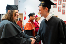 On the left is a middle-aged woman wearing a black toga and biretta. On the right, a young man in a black toga. They are holding hands in a gesture of congratulations. They smile, looking directly at each other. The dean can be seen in the background.